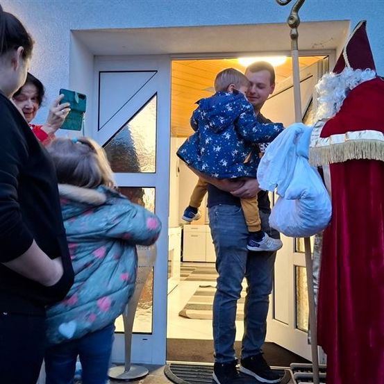 Ein Mann, der ein junges Kind in einem blauen Mantel hält, steht vor einer Tür. Hinter ihm hält ein weiterer Mann, der als Weihnachtsmann verkleidet ist, einen Beutel. Zwei Frauen und ein Mädchen beobachten, eine macht ein Foto.