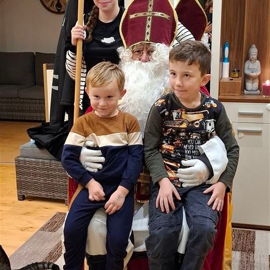 Zwei junge Jungen sitzen auf dem Schoß des Weihnachtsmanns, gekleidet in sein traditionelles rot-weißes Outfit. Ein Mädchen steht hinter ihnen und hält einen Stock. Die Umgebung scheint drinnen zu sein, mit einer Couch und einer Uhr im Hintergrund.