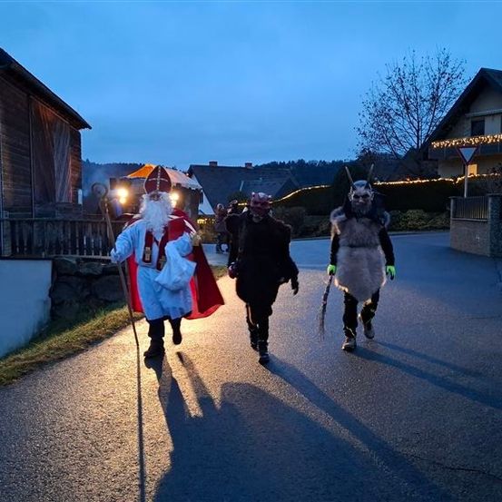 Drei Personen in Kostümen gehen nachts auf einer Straße, eine in einem Weihnachtsmannkostüm und einem roten Umhang, während die anderen Pelzkostüme tragen und Stöcke halten.