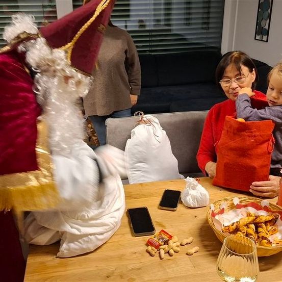 Eine Frau und ein Kind halten einen Geschenkbeutel, während eine als Weihnachtsmann verkleidete Person über einem Tisch gebeugt ist. Ein Sofa und gerahmte Kunst sind im Hintergrund zu sehen.