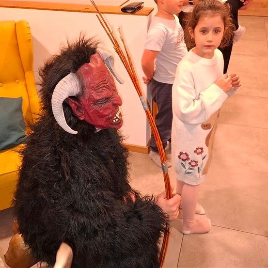 Eine Person in einer Teufelsmaske sitzt auf einem Stuhl neben einem Kind, während ein Junge dahinter steht. Die Szene spielt sich in einem Raum mit gefliestem Boden ab.