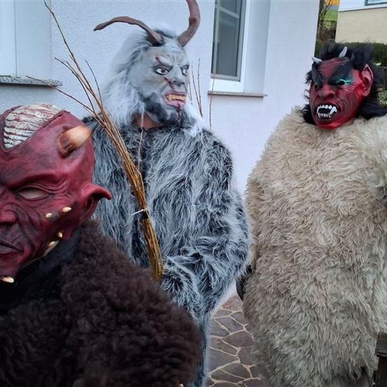 Drei Personen in Kostümen stehen in der Nähe eines Gebäudes. Sie tragen Hörner- und Fangmasken, zwei mit roten Masken und einer mit einem weißen Pelzmantel.