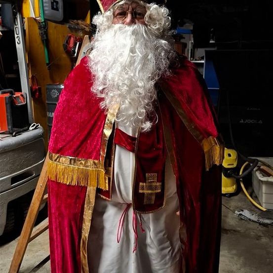 Eine Person, die als Weihnachtsmann verkleidet ist, steht in einer Garage und trägt einen roten Hut und Umhang mit Goldbesatz. Hinter ihm befinden sich Lagerboxen und ein gelber Staubsauger.