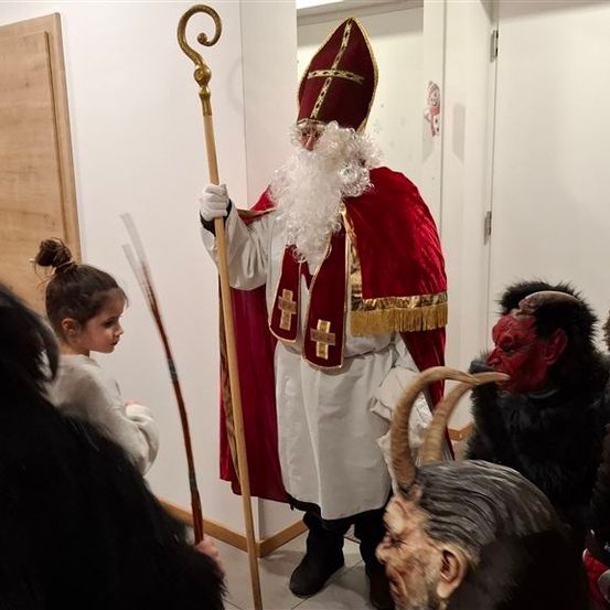 Eine Person in Santa-Claus-Verkleidung steht mit einem Stab, umgeben von Menschen in Dämonenkostümen, von denen einer ein Horn hält. Ein junges Mädchen schaut zu.