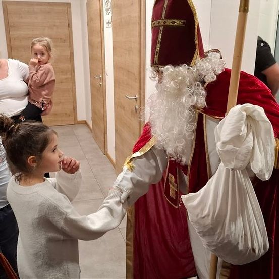 Eine Familie steht in einem Flur, ein junges Mädchen begrüßt einen Mann in Santa Claus-Kostüm. Eine Frau macht ein Selfie, ein Kind ruht auf ihrem Schoß. Hinter ihnen führen Holztüren zu Räumen.