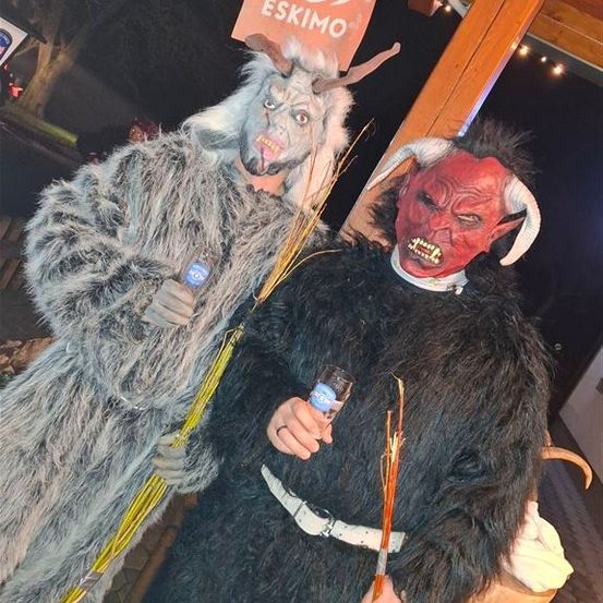 Zwei Personen, die als Dämonen verkleidet sind, posieren nachts für ein Foto, einer hält ein Getränk, beide mit Hörnern und einer mit einer roten Maske. Ein Eskimo-Banner hängt im Hintergrund.