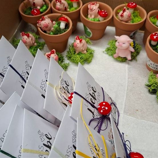 Eine Sammlung kleiner Schweinefiguren mit roten Schleifen, eingebettet in moosgefüllten Töpfen, wird auf einer weißen Oberfläche neben gefalteten weißen Karten mit dem Text 'Liebe' und dekorativen Elementen ausgestellt.