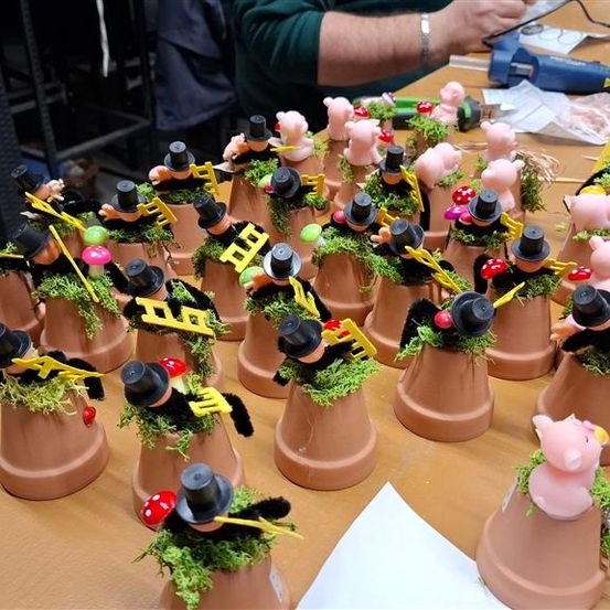 Ein Tisch voller Tongefäße, die mit grünem Moos verziert sind und Miniatur-Schweinefiguren und schwarze Zylinderhüte tragen, wobei einige Schweine gelbe Flügel tragen.