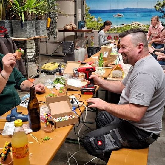 Gruppe von Menschen in einer Werkstatt, zwei Männer stellen etwas mit Kleber und Flaschen her, einer lächelt, Werkzeuge auf dem Tisch, Pflanzen, Meerblick im Hintergrund.