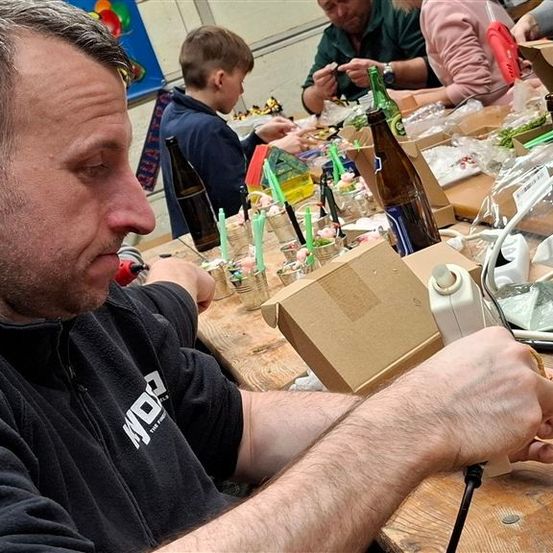 Ein Mann in einer schwarzen Kapuzenjacke arbeitet an einem Projekt, umgeben von verschiedenen Gegenständen wie Flaschen, Boxen und einem Gerät. Andere Personen sitzen am selben Tisch und beschäftigen sich mit ähnlichen Aktivitäten.