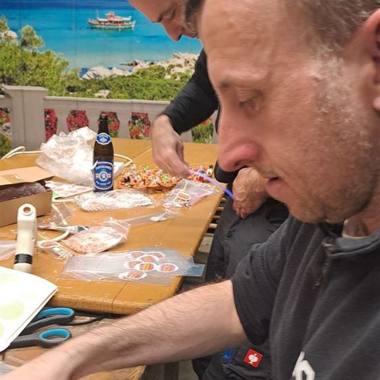 Zwei Männer sitzen an einem Tisch, einer von ihnen schaut auf ein Stück Papier. Eine Bierflasche und verschiedene Gegenstände liegen auf dem Tisch.