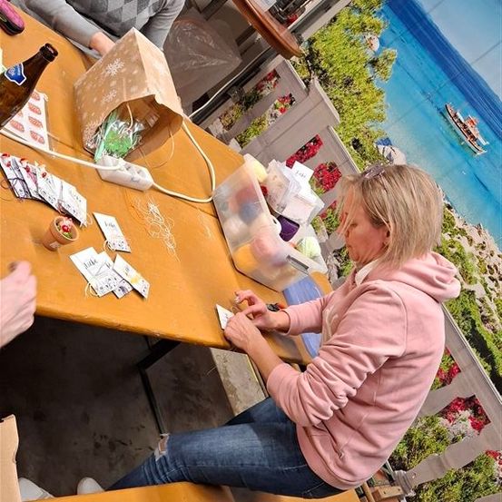 Zwei Frauen sitzen auf einer Bank und basteln mit verschiedenen Gegenständen auf einem Tisch, einer Flasche und einer malerischen Kulisse.