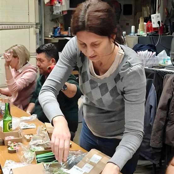 Drei Personen befinden sich in einem Raum. Eine Frau steht und scheint etwas auf dem Tisch zu tun. Der Tisch hat Flaschen, Boxen und andere Gegenstände. Zwei andere Personen sitzen hinter dem Tisch.