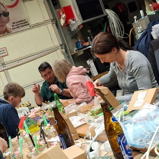 Mehrere Personen sitzen um einen Tisch herum, möglicherweise in einer Garage, und verpacken Gegenstände in Kisten. Flaschen, Becher und andere Gegenstände stehen auf dem Tisch. Eine Frau in einem grauen T-Shirt schaut in die Kiste, die sie verpackt. Dahinter befindet sich ein Regal mit Gegenständen, einem Feuerlöscher und einem Poster mit einem Bild einer Frau.