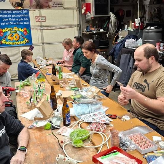 Eine Gruppe von Menschen versammelt sich um einen langen Tisch in einer Garage, mit Flaschen, Werkzeugen und einem Banner. Das Banner lautet 'Oh Weh, Oh Schreck, Schon 40 Jahr'. Die Leute scheinen an einem Projekt zu arbeiten.