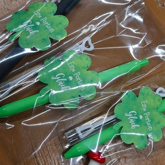 Mehrere grüne Stifte befinden sich in Plastikverpackungen mit Kleeblatt-Designs. Jeder Stift hat einen kleinen roten Verschluss und ein Etikett, das 'Eine Portion Gluck' lautet. Die Verpackung ist transparent, und die Stifte sind auf einer hölzernen Oberfläche angeordnet.
