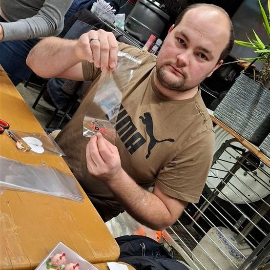 Ein Mann in einem braunen T-Shirt hält eine Plastiktüte in einer Werkstatt. Er betrachtet den Inhalt, während er an einem Holztisch sitzt. In der Nähe steht eine Schachtel mit Schweinefiguren und Scheren. Im Hintergrund sitzt auch eine Frau.