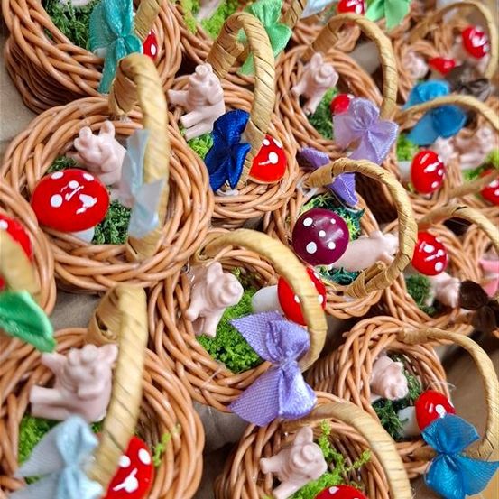 Viele kleine Weidenkörbchen sind mit Schweinefiguren, Moos und bunten Schleifen gefüllt. Die Körbchen sind in einer Reihe angeordnet, und die Schweine tragen rote Pilzhüte.
