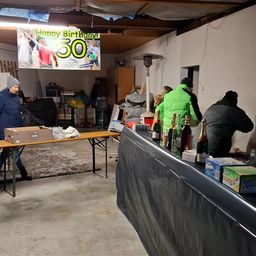 Eine Geburtstagsfeier in einer Garage mit einem Tisch, Flaschen und einem Banner. Menschen stehen herum.
