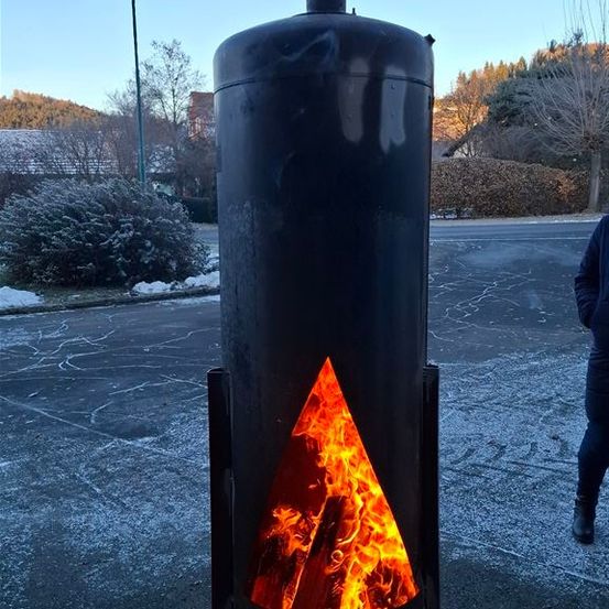 Ein großer schwarzer zylindrischer Ofen mit einem brennenden Feuer darin, draußen in einem verschneiten Bereich, mit einer Person in der Nähe.
