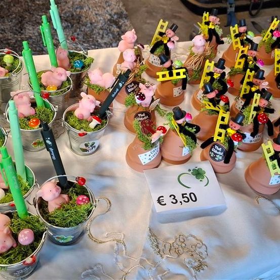 Ein Tisch ist mit einem weißen Tuch bedeckt und hat mehrere kleine Töpfe mit rosa Schweinefiguren, grünem Gras und dekorativen Elementen. Jeder Topf hat einen grünen Stift und einen Preisschild von 3,50 Euro.