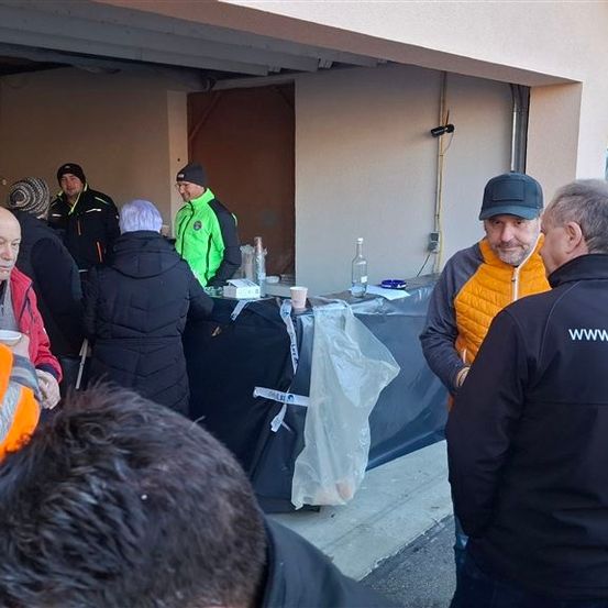 Mehrere Personen in Winterkleidung versammeln sich in einer Garage und sehen einem Tisch mit einem Becher und einer Flasche zu. Ein Mann in einer gelben Weste spricht mit einem anderen Mann.