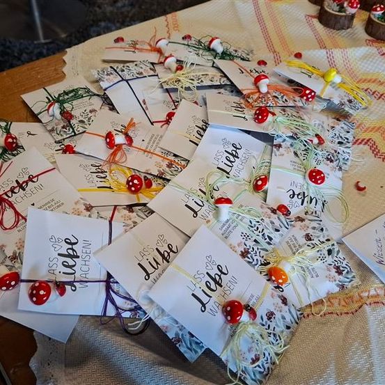 Eine Sammlung personalisierter Geschenke mit Blumen- und Pilzdesigns, verpackt in weißes Papier und mit bunten Bändern gebunden. Der Text 'Liebe' steht auf einigen der Etiketten. Die Gegenstände sind auf einem mit einem weißen Tuch bedeckten Tisch angeordnet.