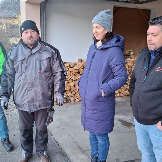 Vier Personen stehen auf einem Parkplatz. Sie tragen Winterkleidung. Hinter ihnen befindet sich eine Wand mit gestapelten Holzscheiten.