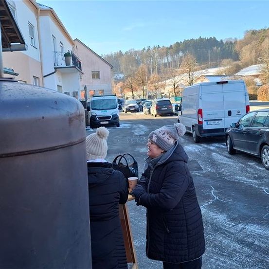 Zwei Frauen in Wintermänteln stehen neben einer großen Metallstruktur, eine hält eine Tasse. Sie stehen vor einem Tisch auf einem verschneiten Parkplatz mit mehreren geparkten Autos und Gebäuden im Hintergrund.