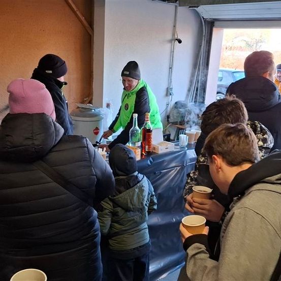 Eine Gruppe von Menschen steht um einen Tisch mit Flaschen, Tassen und einem Fass. Sie sind warm gekleidet, wahrscheinlich in einer kalten Umgebung. Eine Person trägt eine reflektierende Weste.