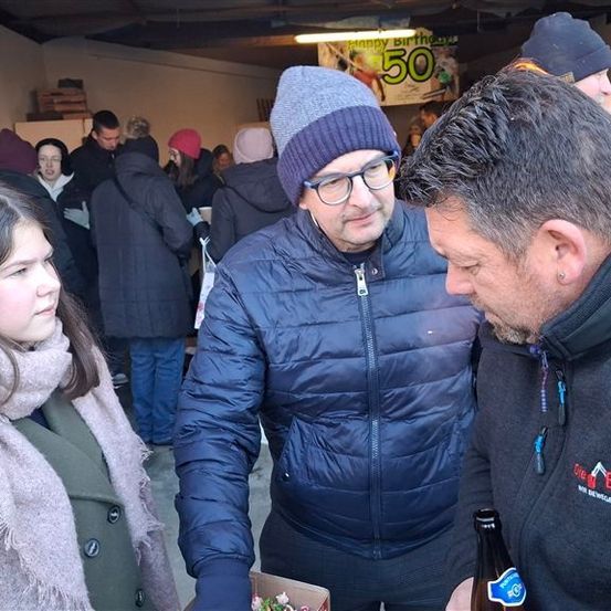 Eine Gruppe von Menschen in Winterkleidung steht in einem Raum. Ein Mann mit Brille hält Blumen, während ein anderer Mann eine Flasche hält. Ein Schild sagt Happy Birthday 50.
