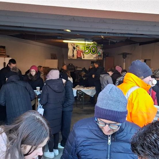 Eine Gruppe von Menschen ist drinnen versammelt, wahrscheinlich bei einer Party. Einige tragen Mützen und Brillen. Ein Tisch mit Tassen ist im Vordergrund.