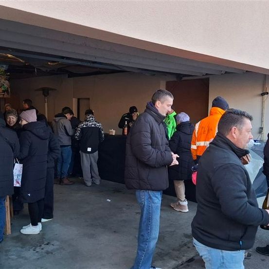 Eine Gruppe von Menschen in Winterkleidung steht in einer Schlange bei einer Feier in einer Garage. Ein Geburtstagsbanner hängt über ihnen.