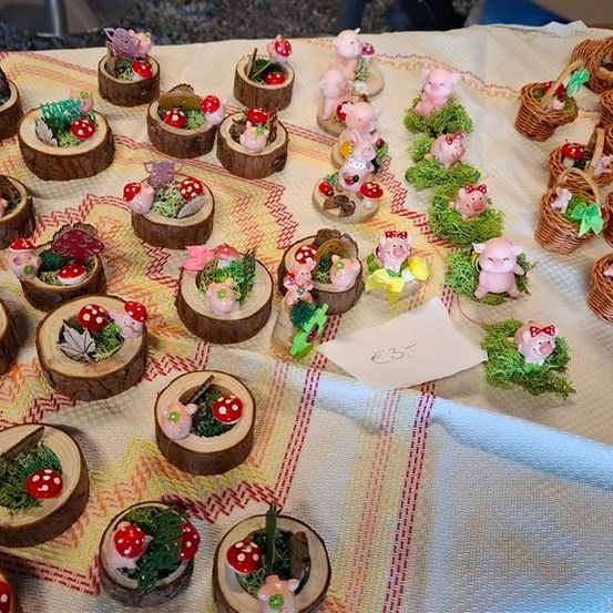 Ein Tisch, der mit einem weißen Tuch bedeckt ist, zeigt eine Vielzahl von Miniatur-Schweinefiguren und kleinen Körben, die jeweils mit Pilzen und Grünzeug verziert sind, zusammen mit einem Zettel, der einen Preis von 35 Euro anzeigt.