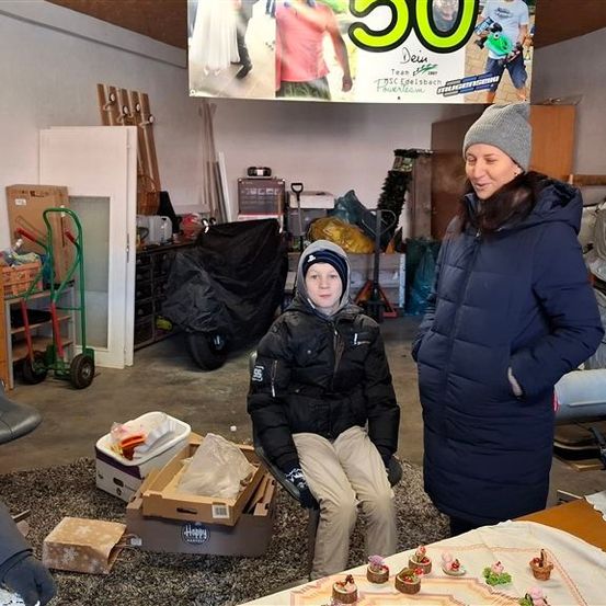 Zwei Frauen und ein Kind stehen in einer überfüllten Garage mit einem Banner, Cupcakes und einem Hintergrund. Ein Schreibtisch mit Gegenständen und ein Stuhl sind in der Nähe.