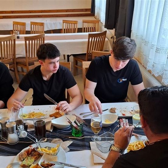 Eine Gruppe von Menschen sitzt in einem Restaurant an einem Tisch und isst eine Mahlzeit mit Tellern, Gläsern und Besteck. Sie tragen schwarze T-Shirts. Eine Person hält ein Messer. Leere Stühle umgeben sie.