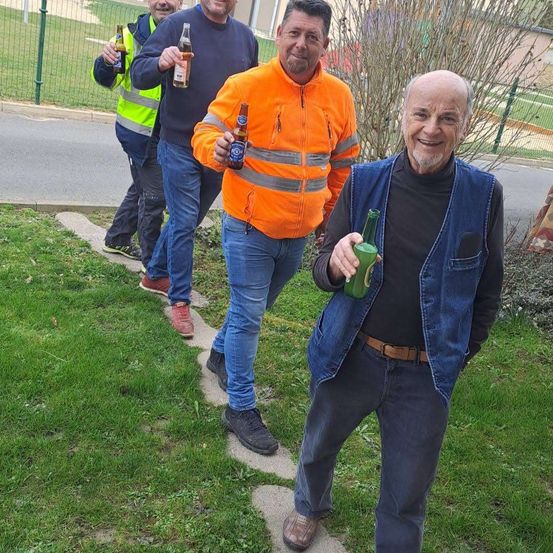Vier Männer stehen auf einem Bürgersteig, einer mit einer grünen Flasche, zwei mit braunen Flaschen. Sie lächeln, und einer trägt eine reflektierende Weste.