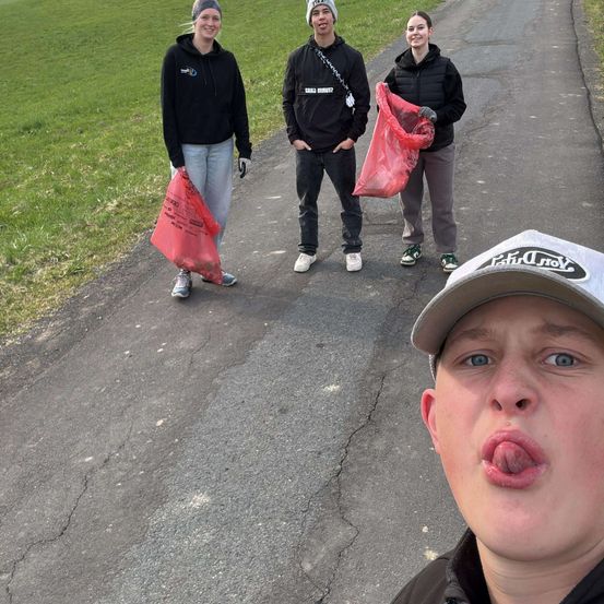 Drei Personen in schwarzen Hoodies und Hosen sammeln Müll auf einer Straße mit einem Grasfeld an der Seite.