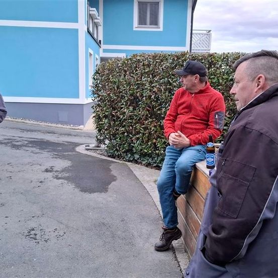 Drei Menschen stehen und sitzen draußen. Eine Frau steht in Jeans und einer grauen Jacke, ein Mann sitzt mit einer Bierflasche auf einer Holzmauer.