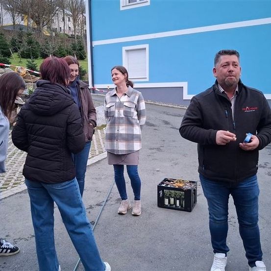 Eine Gruppe von Menschen steht vor einem blauen Haus, zwei Frauen lächeln, ein Mann hält eine kleine Kiste und ein Mann redet.