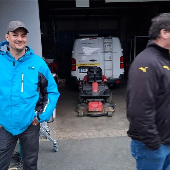 Zwei Männer stehen in einer Garage. Der Mann auf der linken Seite trägt einen blauen Mantel und lächelt in die Kamera. Hinter ihm steht ein weißer Lieferwagen mit einer Leiter an der Seite.