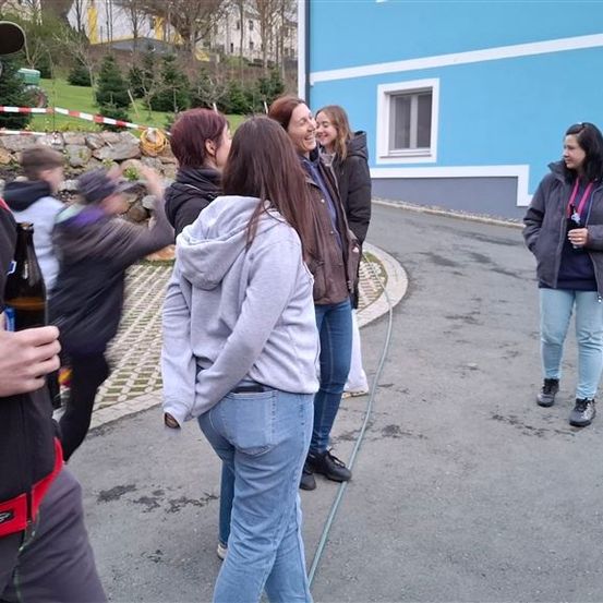 Eine Gruppe von Menschen versammelt sich vor einem blauen Gebäude. Einige lächeln und schauen sich gegenseitig an. Der Mann im Vordergrund hält eine Flasche.