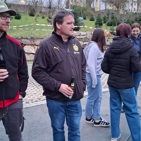 Eine Gruppe von Menschen steht in einem Außenbereich, einige halten Flaschen. Ein Mann in einer Kappe lächelt zur Kamera. Dahinter eine Steinmauer und ein Feld mit Bäumen.