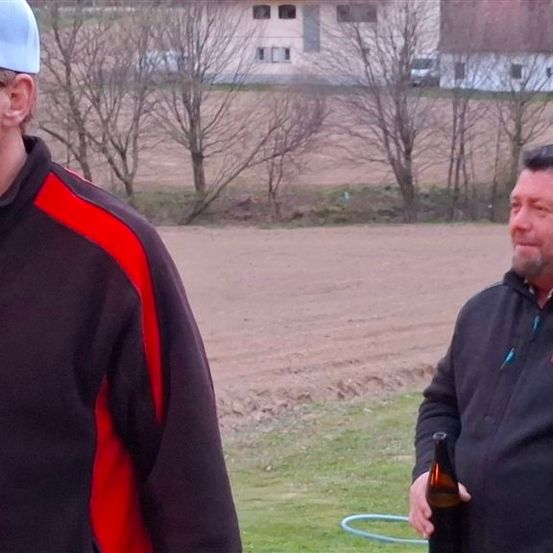 Zwei Männer stehen im Freien, einer hält eine Bierflasche in der Hand, vor einem Haus mit Bäumen und einem Feld.