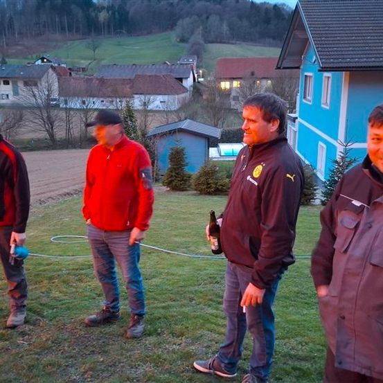Vier Männer stehen auf einem Grasfeld mit einem blauen Haus dahinter. Sie tragen alle Jacken und scheinen ein Gespräch zu führen. Der Mann in der Mitte hält eine Flasche.