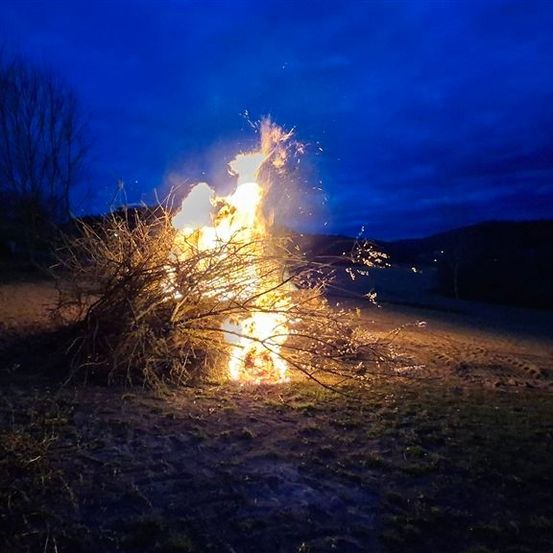 Ein Lagerfeuer brennt bei Nacht in einem Feld mit klarem blauem Himmel. Bäume und Berge sind im Hintergrund.
