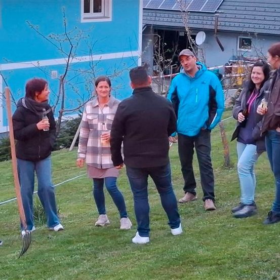 Eine Gruppe von Menschen in lässiger Kleidung steht auf einem Rasen und unterhält sich, während sie Getränke in den Händen halten. Hinter ihnen befindet sich ein Haus mit Solarpanelen und einem Baum.