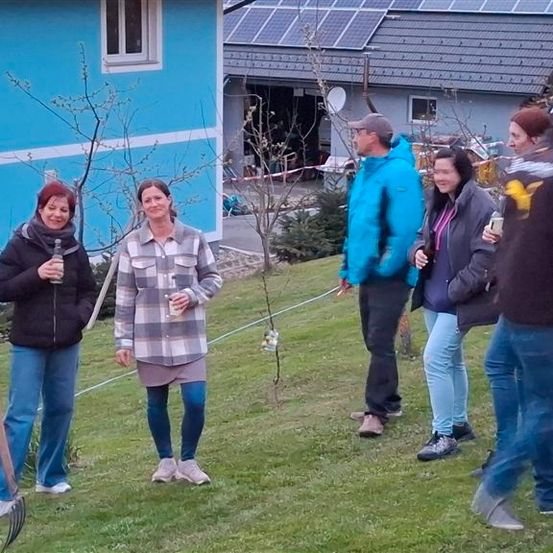 Eine Gruppe von Menschen steht draußen in der Nähe eines Hauses, einige halten Getränke. Eine Frau hält eine Harke. Bäume und ein Haus mit Solarpanelen sind im Hintergrund.
