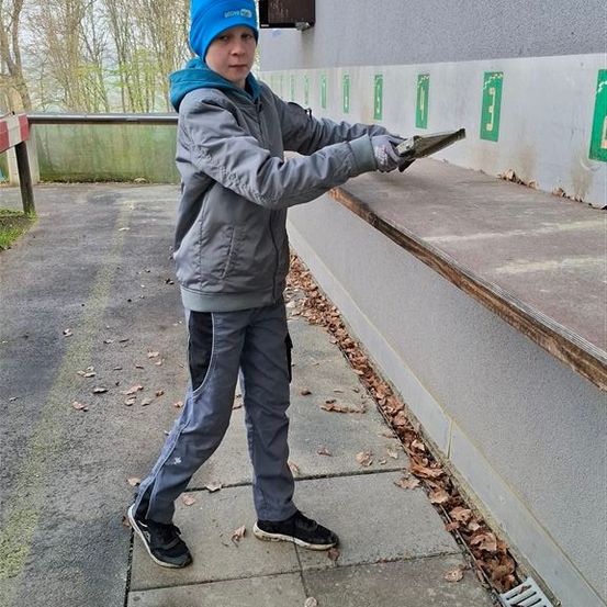 Ein Junge mit einer blauen Mütze und einem grauen Jackenmantel hält eine Steinplatte, möglicherweise zur Reparatur einer Wand. Ein Schild zeigt 'diesen Weg.'