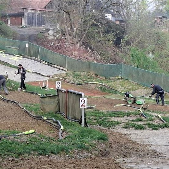 Drei Arbeiter bereiten eine mit den Nummern 2 und 3 markierte Erdstrecke vor. Die Strecke ist auf einer Seite eingezäunt, und im Hintergrund befindet sich ein Haus mit einer Einfahrt.